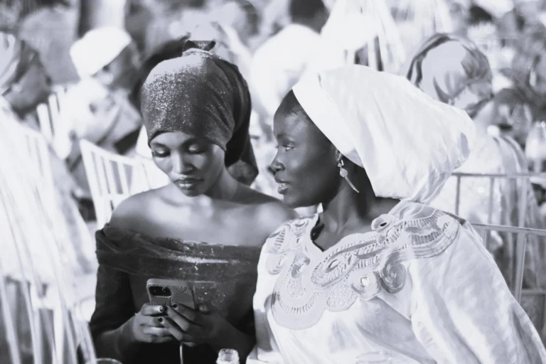 Two women in headwraps lean together to look at a phone in a crowd.