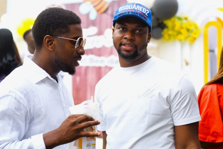 Two men in white tops chat closely outdoors, one wearing a blue cap and holding a drink.