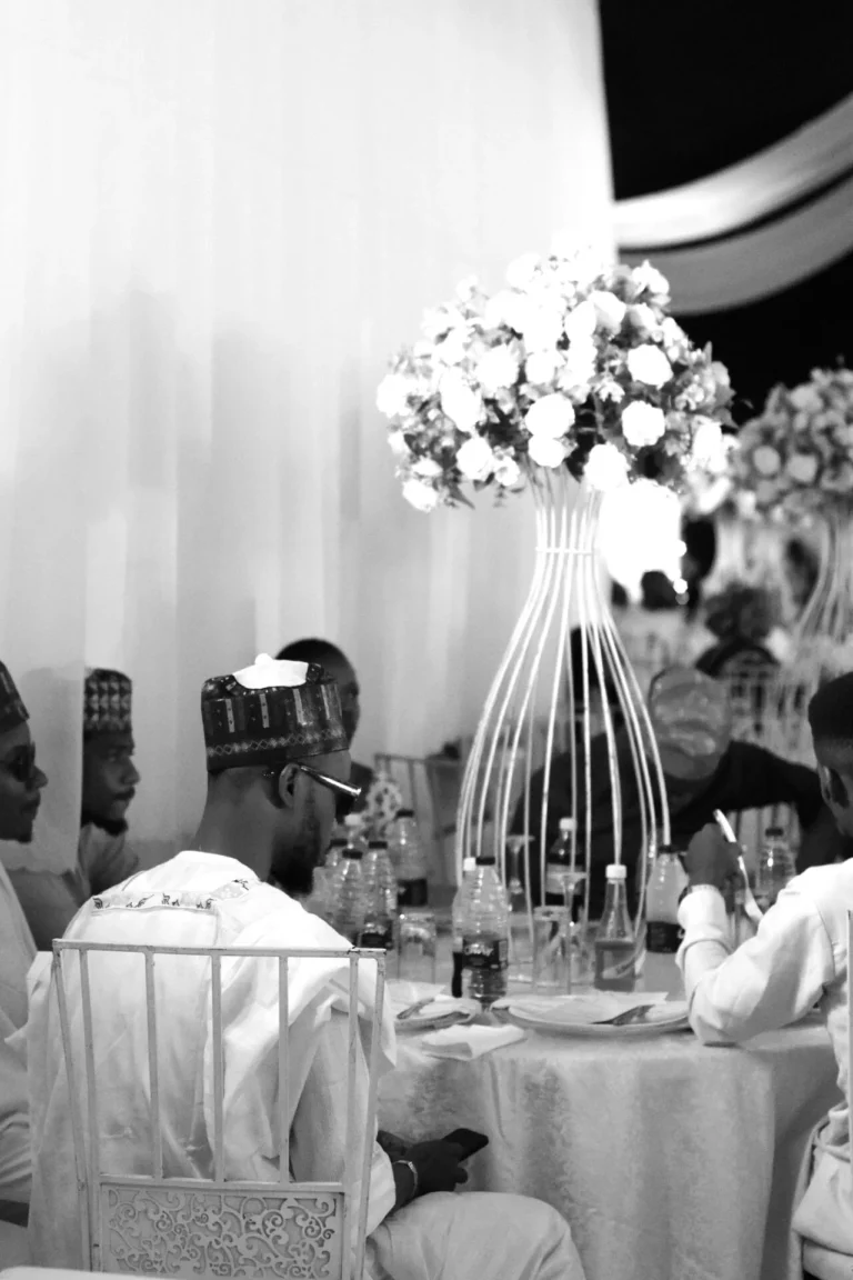 Seated guest in embroidered cap at a reception table with tall floral centrepiece.