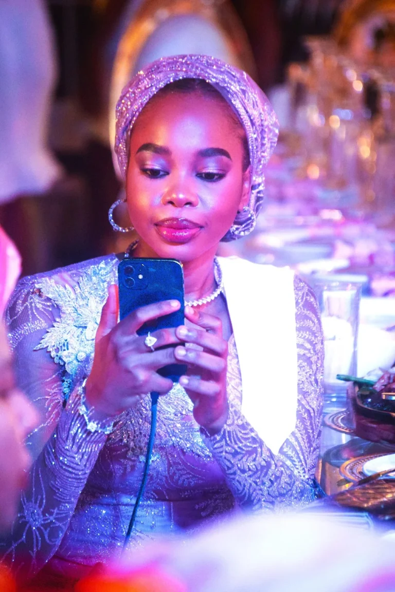 Woman in beaded gown and silver gele takes a selfie under pink ambient lights.