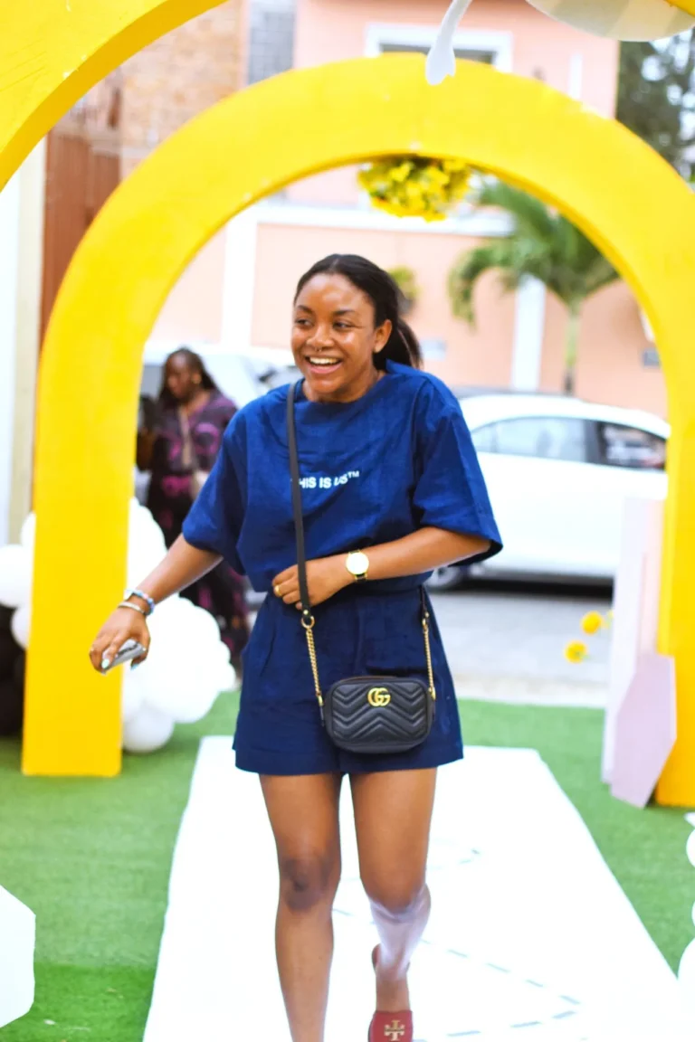 Smiling woman in navy outfit walks through a bright yellow arch onto a white runway carpet.