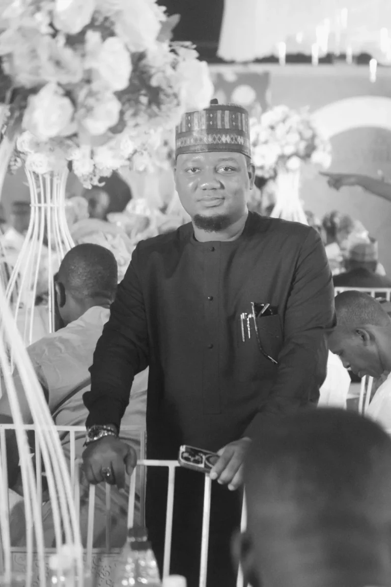 Smiling man in embroidered cap and dark agbada stands beside floral arrangements.