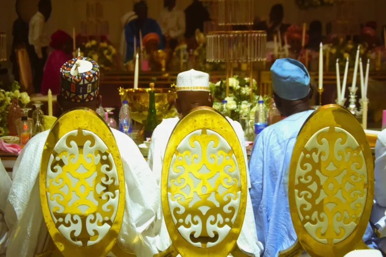 Three men in patterned caps sit on ornate gold-backed chairs at a banquet table.