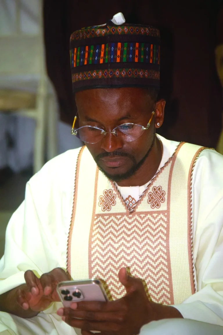 Man in colourful embroidered cap and cream agbada looks down at his phone.