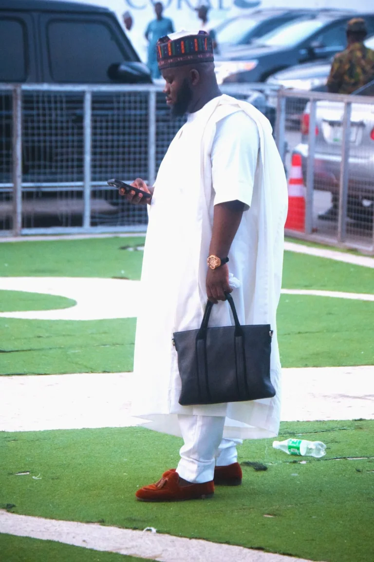 Man in white agbada and patterned cap stands on green astroturf holding a tote bag.