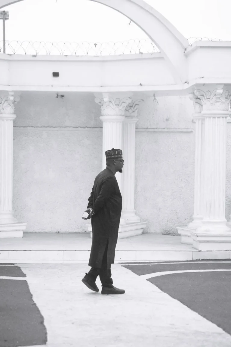 Man in dark agbada walks across paved terrace beneath white classical columns.
