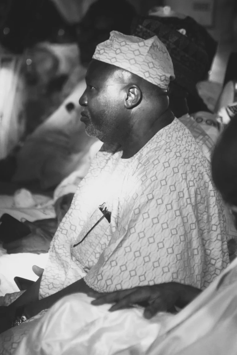 Bearded man in patterned cap and white embroidered robe sits in quiet contemplation.