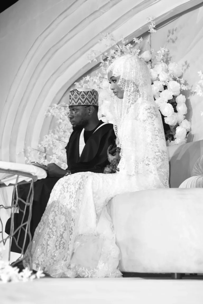 Bride in lace veil and groom in dark robe sit on stage before floral backdrop.