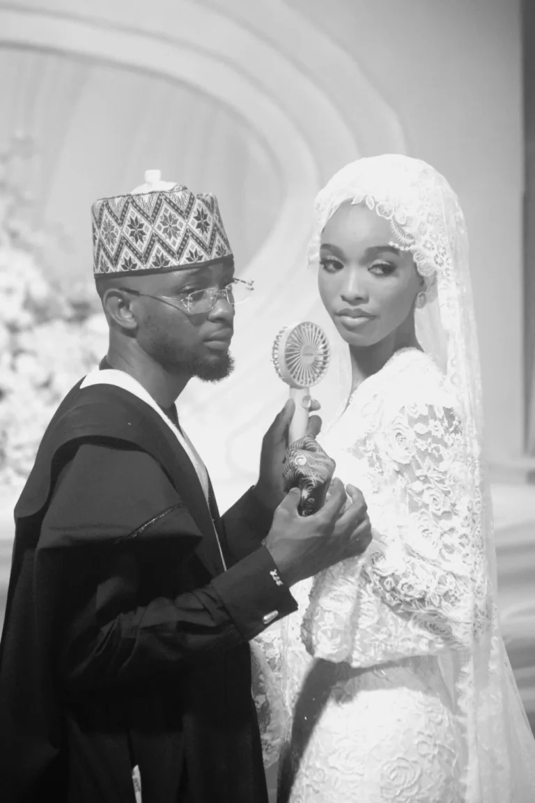 Groom in embroidered cap holds a handheld fan toward bride in lace veil.