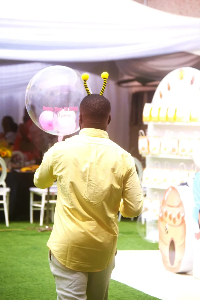 Man in yellow shirt and bee antennae headband holds a Happy First Birthday balloon inside a marquee.