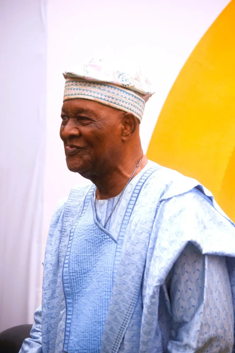 Smiling elderly man wearing a traditional embroidered light blue agbada and matching Yoruba cap.